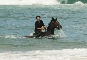 Dressage trainer Maggie Broekman rides a client's horse thigh-deep in ocean surf on New Zealand beach.