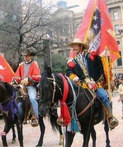 Two mustang horses ridden by colorfully-clad riders carrying flags.