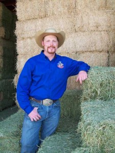 Manager Wayne Rutherford poses in a bright blue ensemble and cowboy hat in front of some bales of hay.