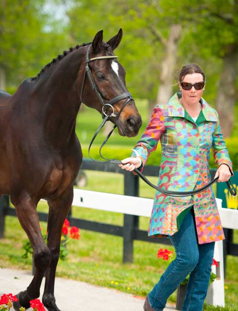 Woman in a brightly-colored coat of geometric pattern leads a bay horse.