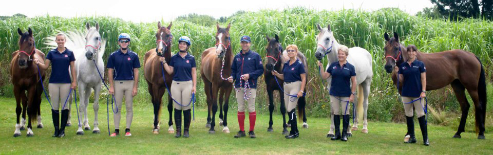 The USA World Endurance Championship team stands with their unsaddled horses before an orchard of greenery.