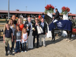 Group of people hold trophy next to rider on blanketed horse.