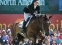 Beezie Madden and her horse Simon clear a jump with the giant Spruce Meadows North American tournament banner and a crowd of people in the backgroud.