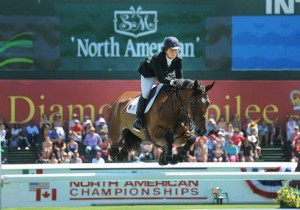 Beezie Madden and her horse Simon clear a jump with the giant Spruce Meadows North American tournament banner and a crowd of people in the backgroud.